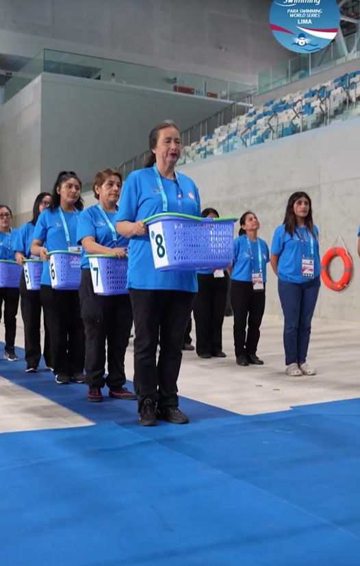 Voluntarias y voluntarios con polos azules llevan las bandejas de medallas en fila junto a la piscina